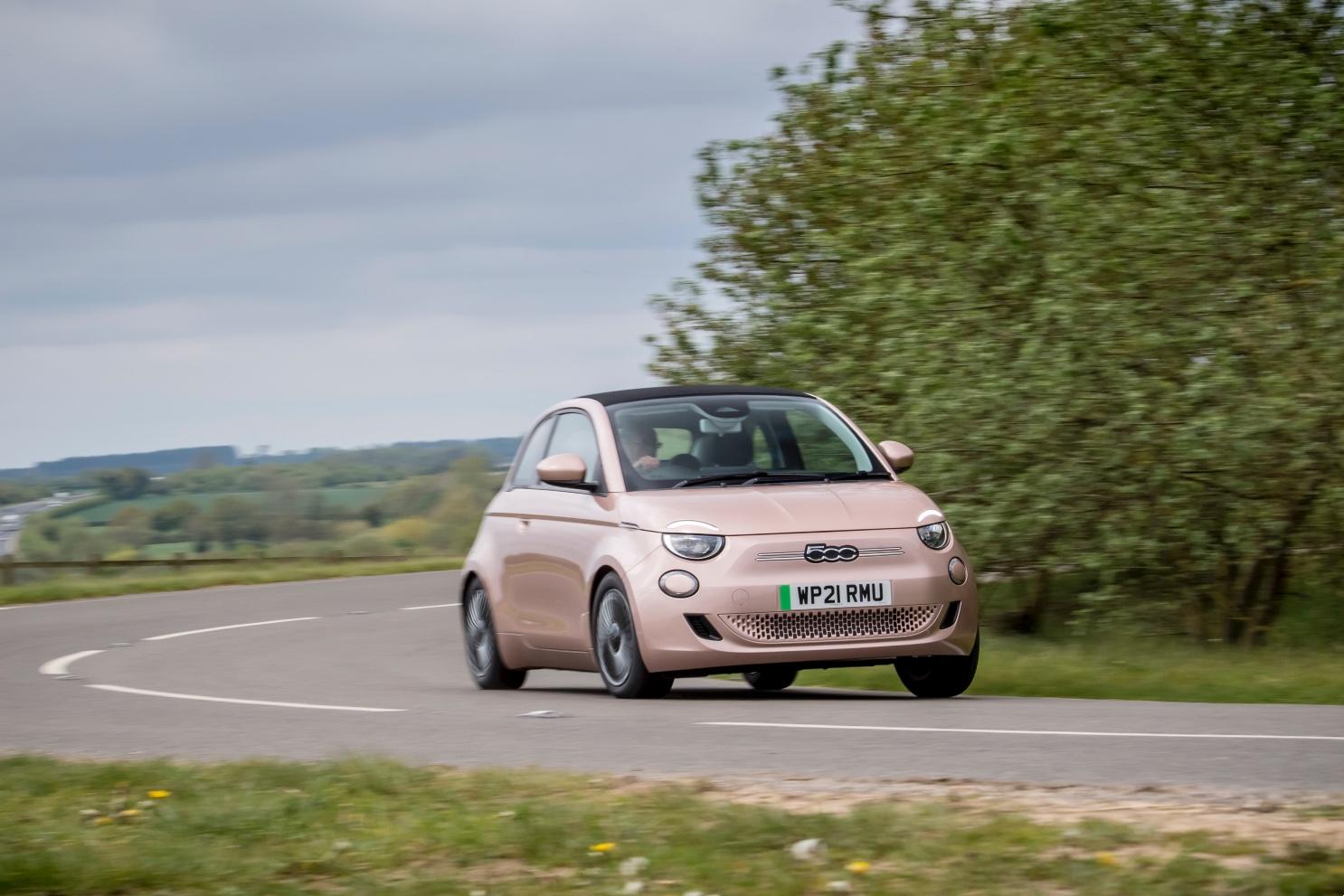 Pink Fiat 500 driving on a curved rural road, surrounded by greenery and a cloudy sky.
