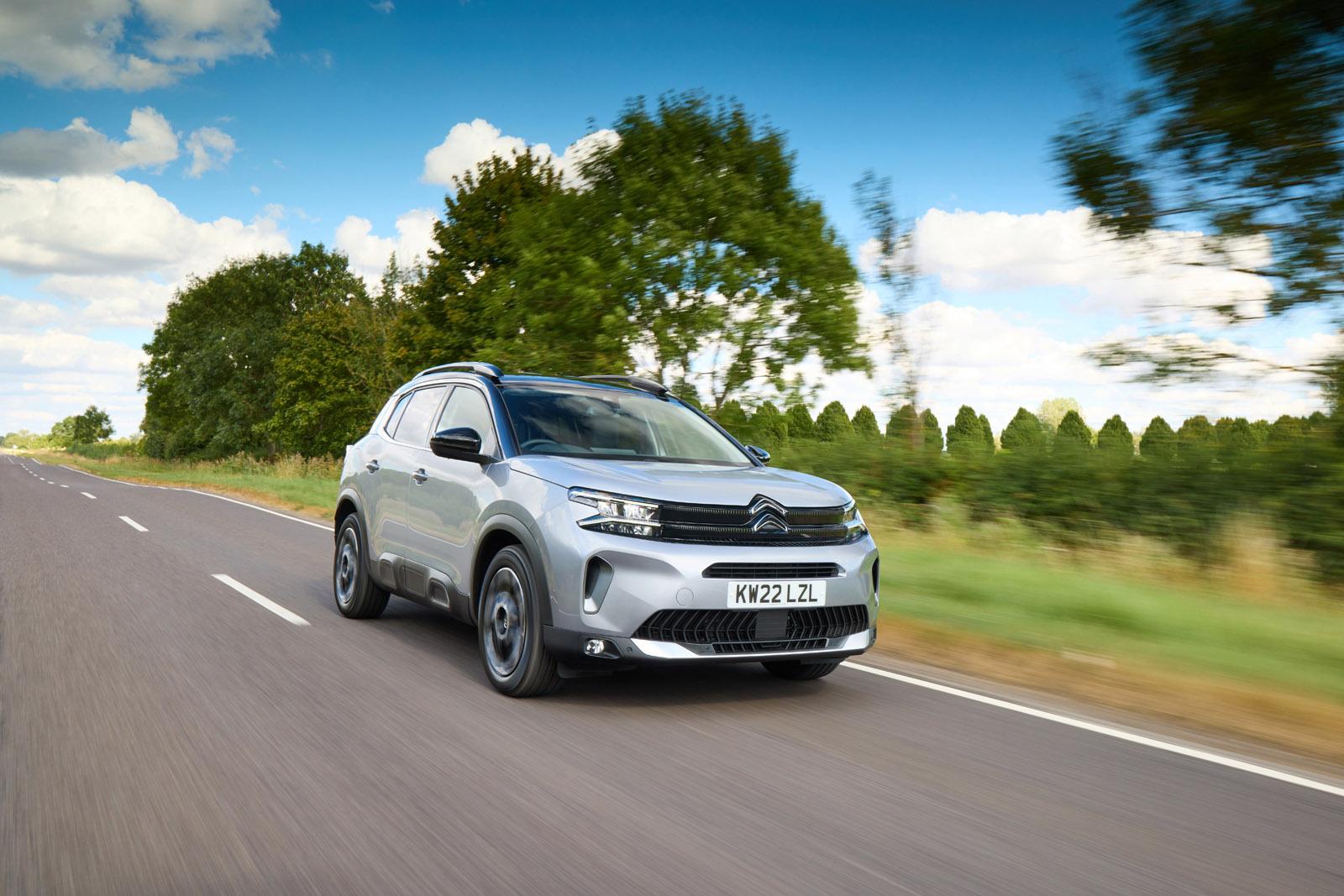 A silver SUV driving on a rural road with blurred trees and a blue sky with clouds in the background.