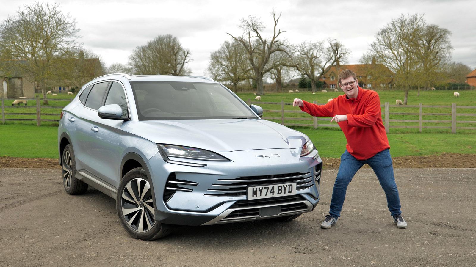 A person in a red sweater poses excitedly next to a silver SUV parked on a rural road with fields and trees in the background.