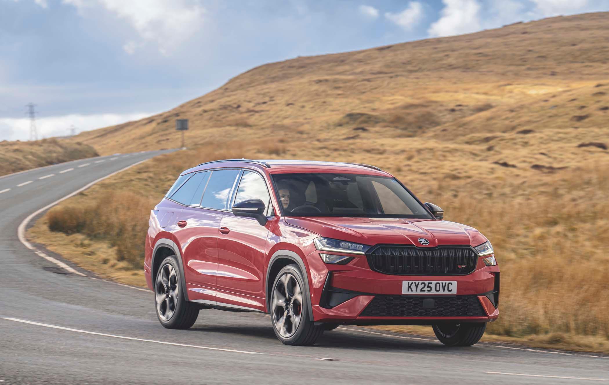 Red Škoda Kodiaq driving on a winding road through a hilly landscape under a partly cloudy sky.