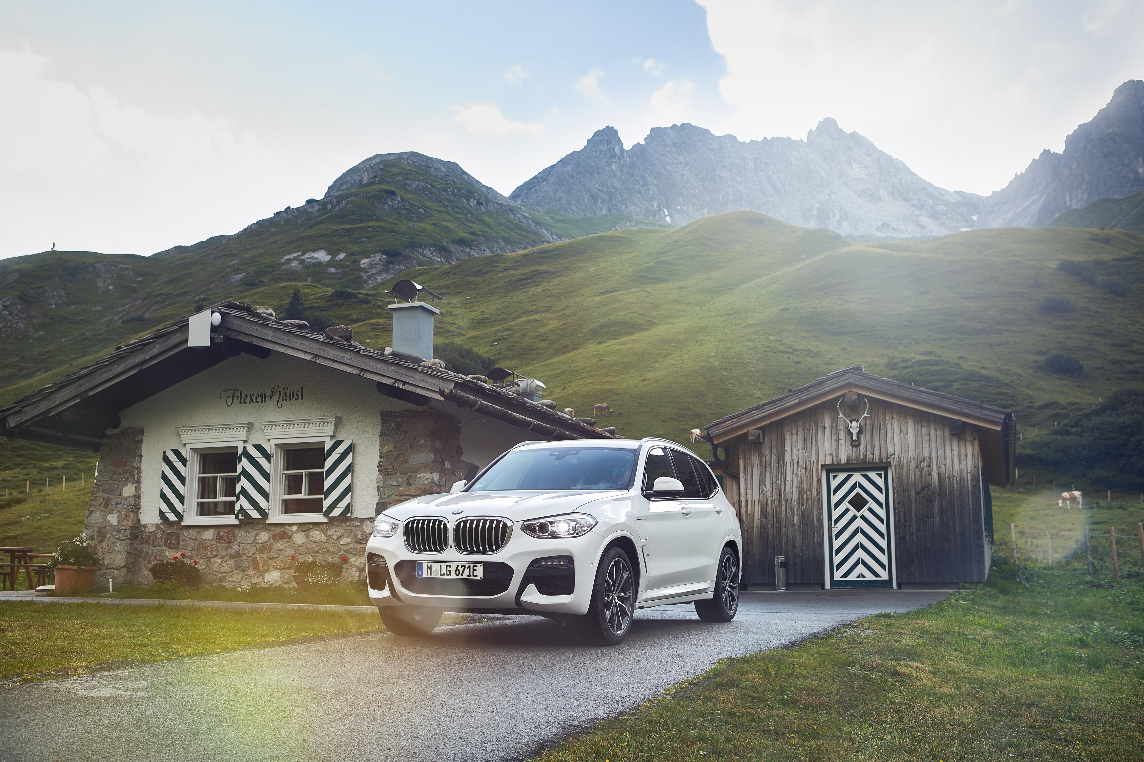 a white BMW X3 hybrid parked outside a small wooden house in the mountains