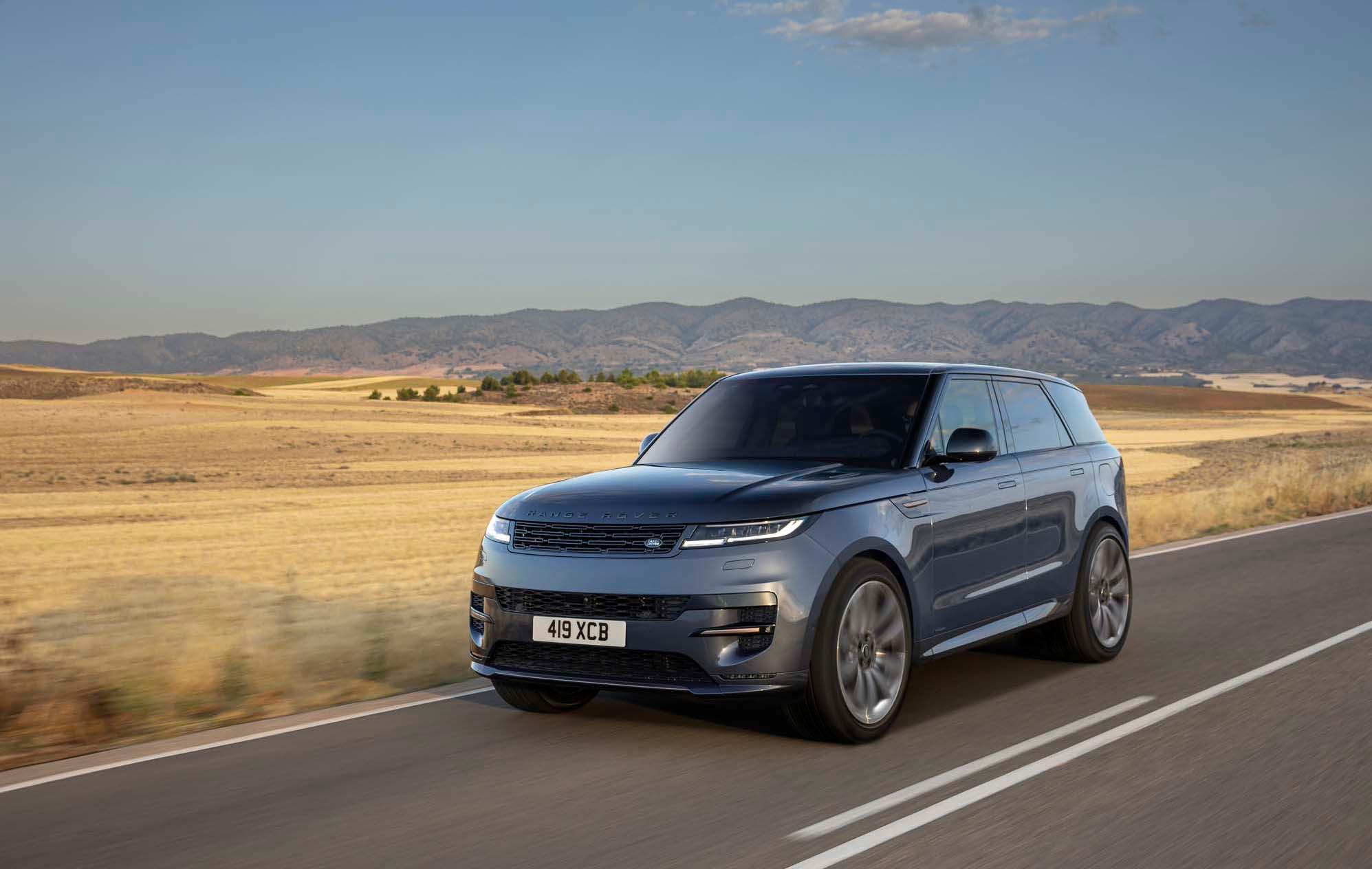 A dark grey Range Rover Sport drives on a rural road with vast fields and distant mountains under a clear blue sky.